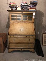 Full front view of solid wood secretary desk with books on top, fold-out writing surface closed, and visible two large drawers at base.