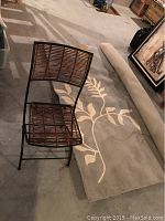 Photo showing the folded iron and reed chair placed on the gray floral area rug with part of the rug rolled up beside it.