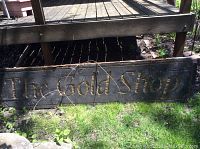 Large wooden sign with gold lettering reading 'The Gold Shop' lying on grass under deck