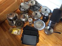Photo of various pots, pans, kettle, waffle maker plates, coffee press, garbage can, and moka pot arranged on wooden floor.