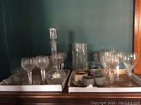 Wide view of assortment of glassware on two trays including wine glasses, decanter, vase, and other glass items