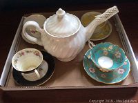 White ceramic teapot with three tea cups and two saucers arranged in a box. Two tea cups have floral designs; one is black edged, the other turquoise with gold and pink flowers.
