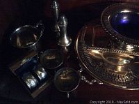 Photo showing assorted silver plated items including salt and pepper shakers, napkin rings in case, and small serving pieces on dark surface.