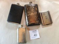 Set of three metal flasks and one metal cigarette holder arranged on white background with lot card included.