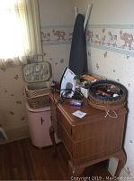 Sewing machine table setup with iron on top, sewing supply baskets, pink hamper, and ironing board against wall with wallpaper.
