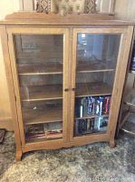 Front view of wooden display cabinet with two glass doors and three interior wood shelves. Some items like books and DVDs are on lower shelves.