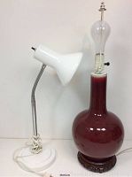 Both lamps shown side by side on a white surface. The white desk lamp has a round base and flexible chrome neck with a flared conical shade. The red ceramic lamp is tall with a rounded body on a carved wooden base and a harp with paint wear.