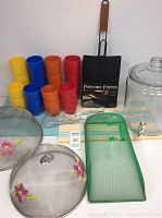 Photo of the entire lot showing six yellow, blue, orange, and red plastic glasses, two flower-patterned metal mesh food covers, a green mesh food cover, the popcorn popper, and the placemats.