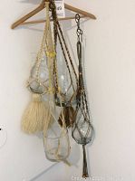 Three vintage macrame plant holders hanging on a wooden hanger against a white wall, each with a clear glass container and tassels.