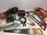 Overall photo of lot showing battery charger, vice, several extension cords, and three saws including one with a wooden handle and one small saw with pin damage