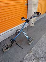 Full view of the assembled Tour Trek collapsible golf push cart with three wheels, blue metal frame, cup holders, and a storage tray on the handle
