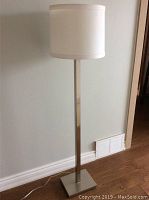 Full view of the floor lamp showing tall silver metal base and square base plate with white fabric drum shade on top.