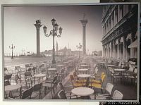 Front view of large framed printed canvas showing St. Marks Square with café tables and chairs, Venetian lamps, columns, and water.