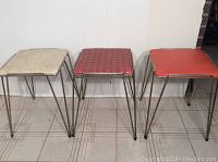 Three vintage stools in a row showing square vinyl seats in white speckled, red speckled, and orange, on tubular metal frames with hairpin legs. The stools are placed on tiled floor.