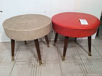 Photo of two vintage circular foot stools, one tan fabric and one red vinyl, showing overall condition and leg details.