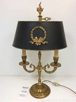 Front view of vintage brass table lamp with black decorative shade, showing dual candle-style bulbs and ornate base.
