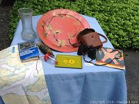 Overview image showing the full collection laid out on a light blue cloth with greenery background, including glass vase, metal tray, ceramic plate, hose coupling, binoculars with leather case, railroad timetable, map, and marine chart.