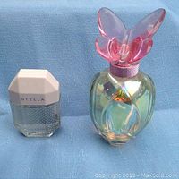 Two perfume bottles on blue cloth background. Left is Stella McCartney Eau de Toilette with geometric white cap, right is M Lucious Pink by Mariah Carey Eau de Parfum with large iridescent bottle and pink butterfly cap.
