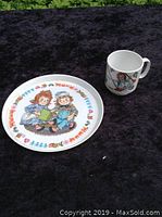 Full view of the Raggedy Ann and Andy child's plate and cup set on a black fabric background