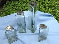 Photo of a set of 4 clear triangular apothecary jars with silver metal lids, displayed outdoors on a blue cloth with greenery in the background.