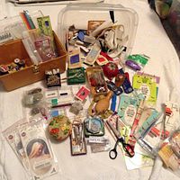 Overview of large sewing lot with scissors, thread, needles, notions, pins, sewing box and more laid out on white surface.