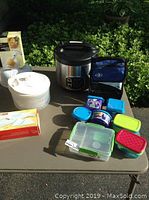 Photo showing all lot items arranged on a table outdoors: rice cooker, salad spinner, lunch containers, and packaged kitchen tools.