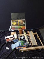 Photo of wooden weaving loom, yarn spools, weaving tools, fabric sample, and instruction booklet arranged on black fabric background.