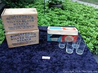 Set of eight clear Pyrex glasses displayed beside original box and two wooden starch boxes on blue velvet surface outdoors.