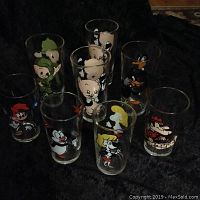 Eight Looney Tunes collector glasses displayed collectively on a dark surface showing characters including Porky Pig, Elmer Fudd, Daffy Duck, Polly, Dudley Do Right, Chilly Willy, Woody Woodpecker.
