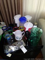 Overview of various pitchers and bar supplies including plastic and ceramic pitchers, glasses with colorful rims, glass shaker, and bar tools in a black tray.