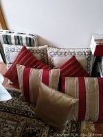 Overview of 7 pillows showing 5 decorative pillows with red, beige, and brown stripes and 2 textured pillows with decorative trim, plus 3 striped lawn chair cushions leaning against the wall