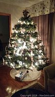 Artificial Christmas tree about 7 feet tall decorated with string lights, ribbons, and ornaments, with white tree skirt beneath.