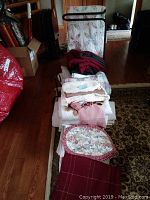 Photo shows a pile of folded linens including a double-sized floral quilt, comforters, towels, and more on a patterned rug. Also visible are multiple towel racks in black and white metal scattered behind the linens.