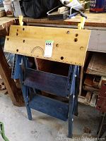 Front view of the portable workbench showing wooden top with holes and yellow adjustment clamps on metal blue frame