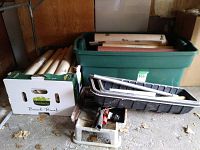 Overall view of a green bin filled with wallpaper rolls and painting supplies, with additional rolls stored in a cardboard box and several painting trays and troughs stacked nearby.