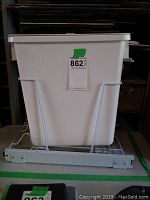 White plastic rectangular garbage can inside a metal wire frame with sliding metal rails on the bottom, shown from front angle.