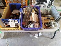 Three containers on a table filled with assorted electrical and hardware items including switches, cover plates, electrical boxes, nuts, bolts, and brackets.