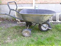 Left side view of a green metal Yardworks wheelbarrow with large bowl, inflated rear tire, and flat front tire showing some surface rust.