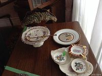 Seven ceramic and china items on a wooden table, including tiered plate with green leafy decoration, floral coasters, maroon ashtrays with gold trim, and small floral dish.