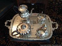 Silver-plated Wm Rogers tea set on an engraved International Silver tray, consisting of teapot, sugar bowl with lid, and creamer, viewed from above on a dark carpet.