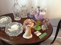 Photo showing various glass and decorative items including a glass decanter, crystal ashtrays shaped with swan designs, brass candlesticks, RCAF paperweight, coasters, and floral decorative pieces on a wooden table.