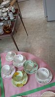 Photo showing six teacups and saucers of various floral patterns laid out on a pink cloth, displaying a variety of pastel colors and gold trim.
