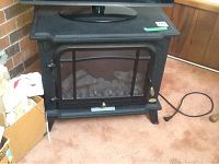Front view of the black Charmglow electric heater showing glass front door with faux logs inside, power cord visible on the carpet.
