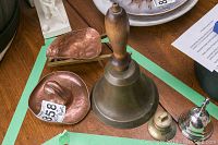 Photo showing the antique bronze school bell with wooden handle alongside two smaller bells and the copper miniatures: a wheelbarrow and a cowboy hat.