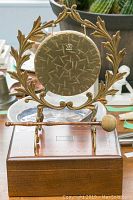 Front view of the brass gong mounted in an ornate leaf-motif brass holder with a wooden mallet resting on the base. The gong surface is textured with a small emblem at the top center.