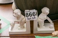 Overall view showing both ceramic dog cricket bookends side by side, each 5x5 inches, with the MaxSold label taped on one bookend.