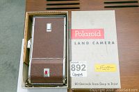 Closed vintage Polaroid Land Camera in original box sleeve, showing brown textured body and silver metal trim, red shutter button.