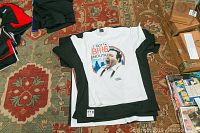 Stack of graphic T-shirts including a white T-shirt with The Honeymooners image and two black T-shirts underneath on a patterned carpet with some boxes nearby.