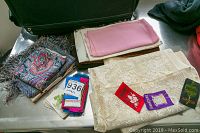 Photo of lace curtains, scarves, and three embroidered badges on a metallic surface beside a suitcase.
