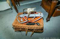 Woven picnic basket topped with an orange extension cord, a silver painting light, and a white corded item, placed on a carpet near wooden furniture.
